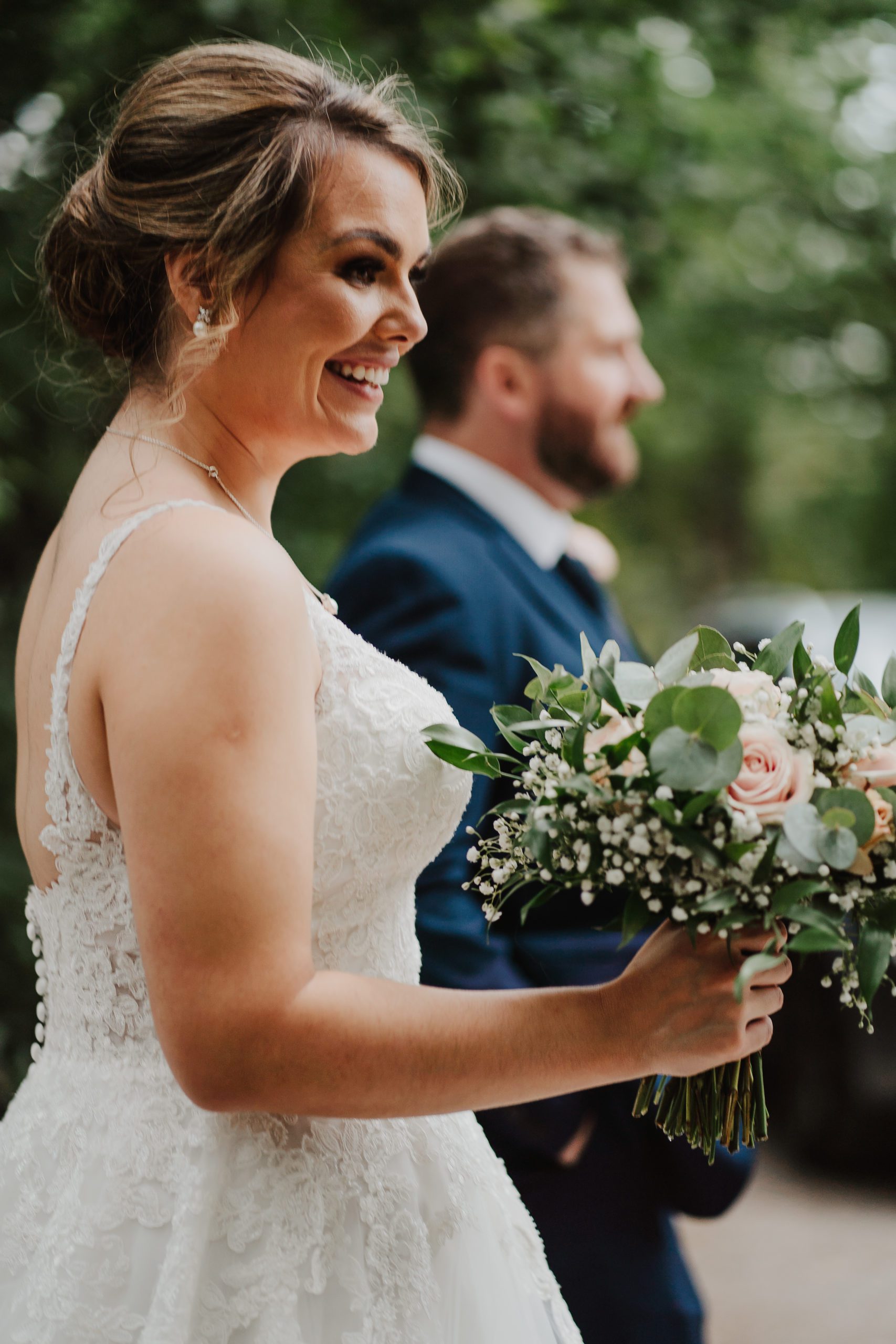 Bride holds bouquet of peachy pink roses smiling with groom out of focus in the background.