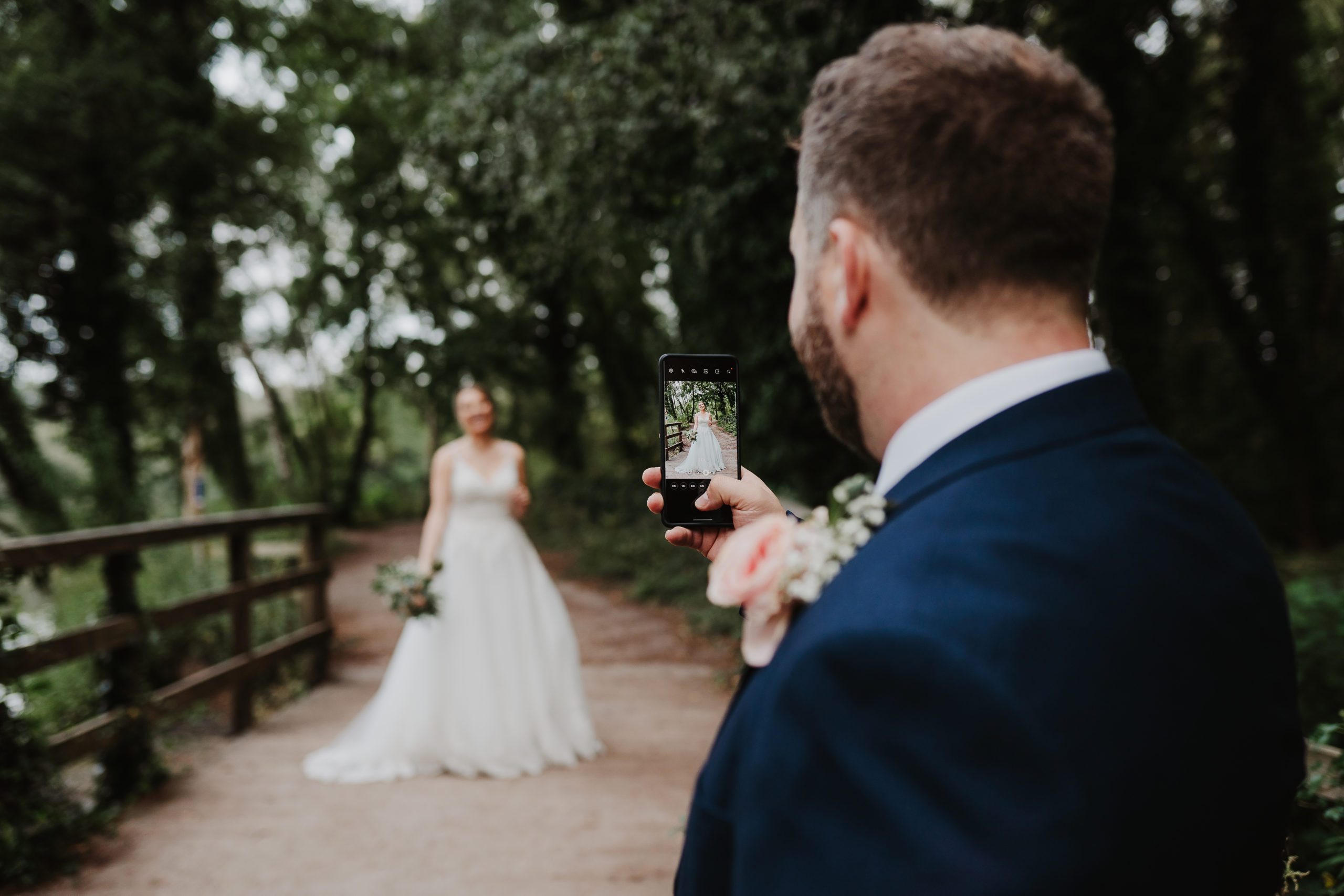 Groom takes photo of bride out of focus in the distance on his mobile phone at Shakerley Mere