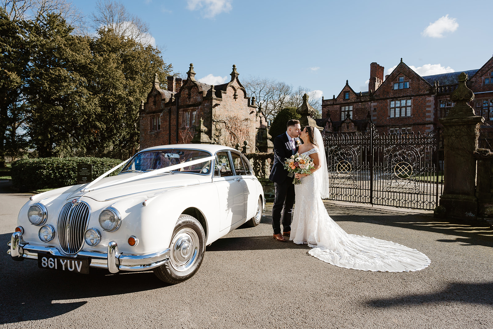 Bride and groom kiss by white Jaguar with Dorfold Hall gates behind