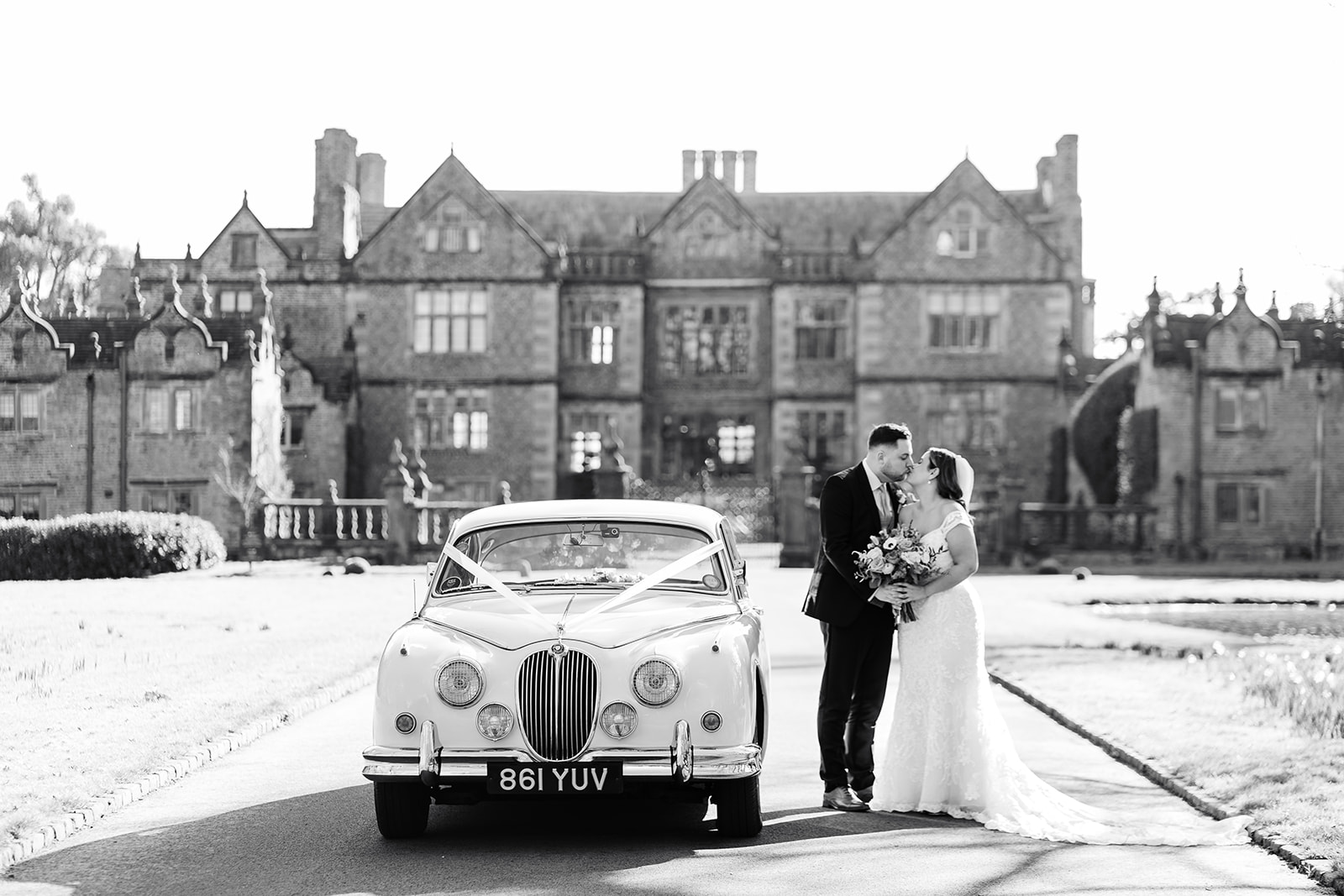 Bride and groom kiss by white Jaguar in front of Dorfold Hall