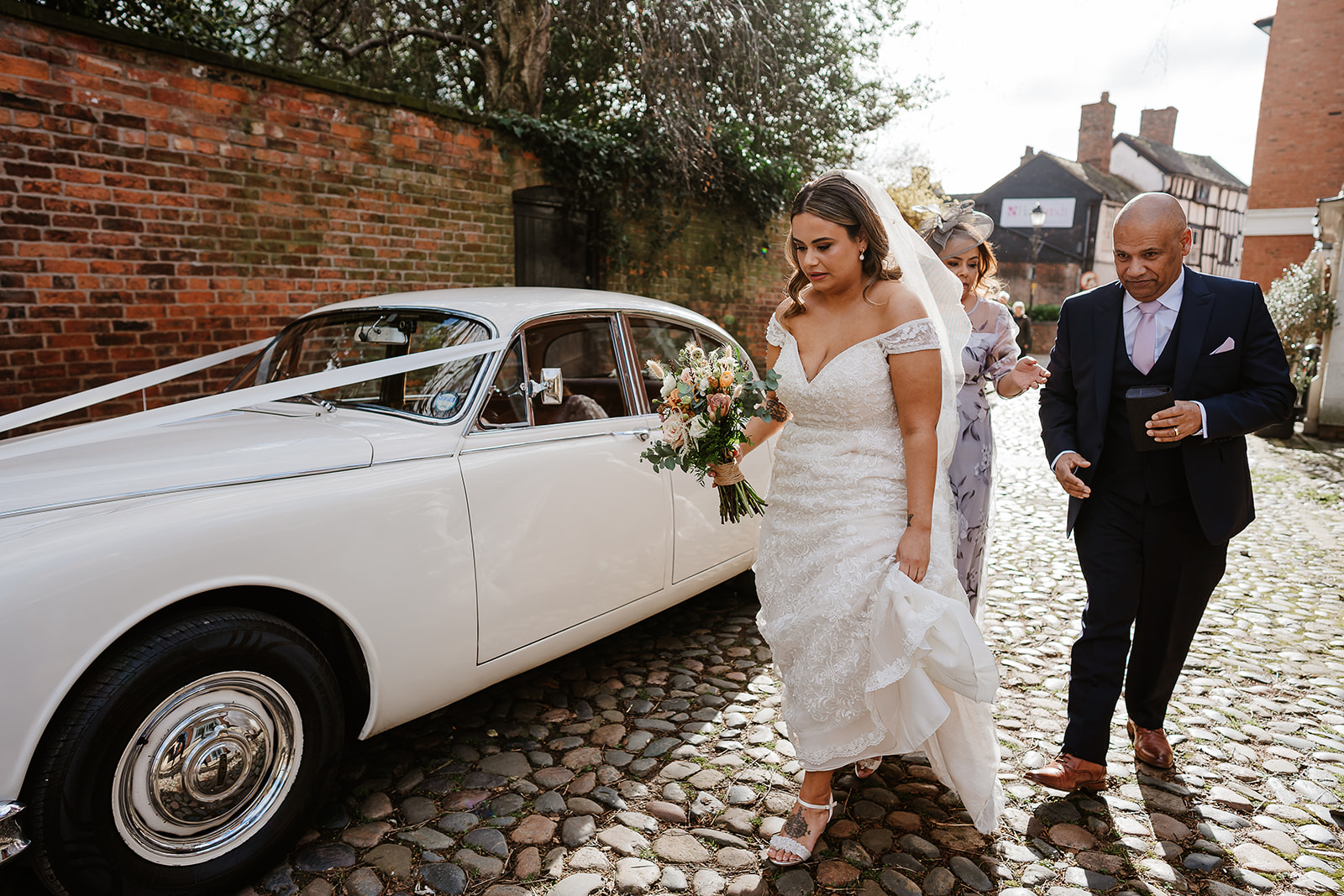 Bride and parents walking on cobbles by white Jaguar wedding car