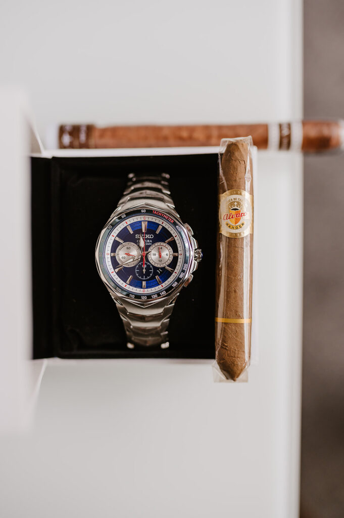 A close-up flat lay image of a seiko watch in it's presentation box.  A cigar in its plastic wrapping sits on top of the presentation box.