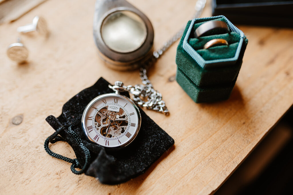 A flat lay of two wedding rings in a racing green velvet ring box sitting alongside cufflinks and a pocket watch sat on top of a black velvet bag.